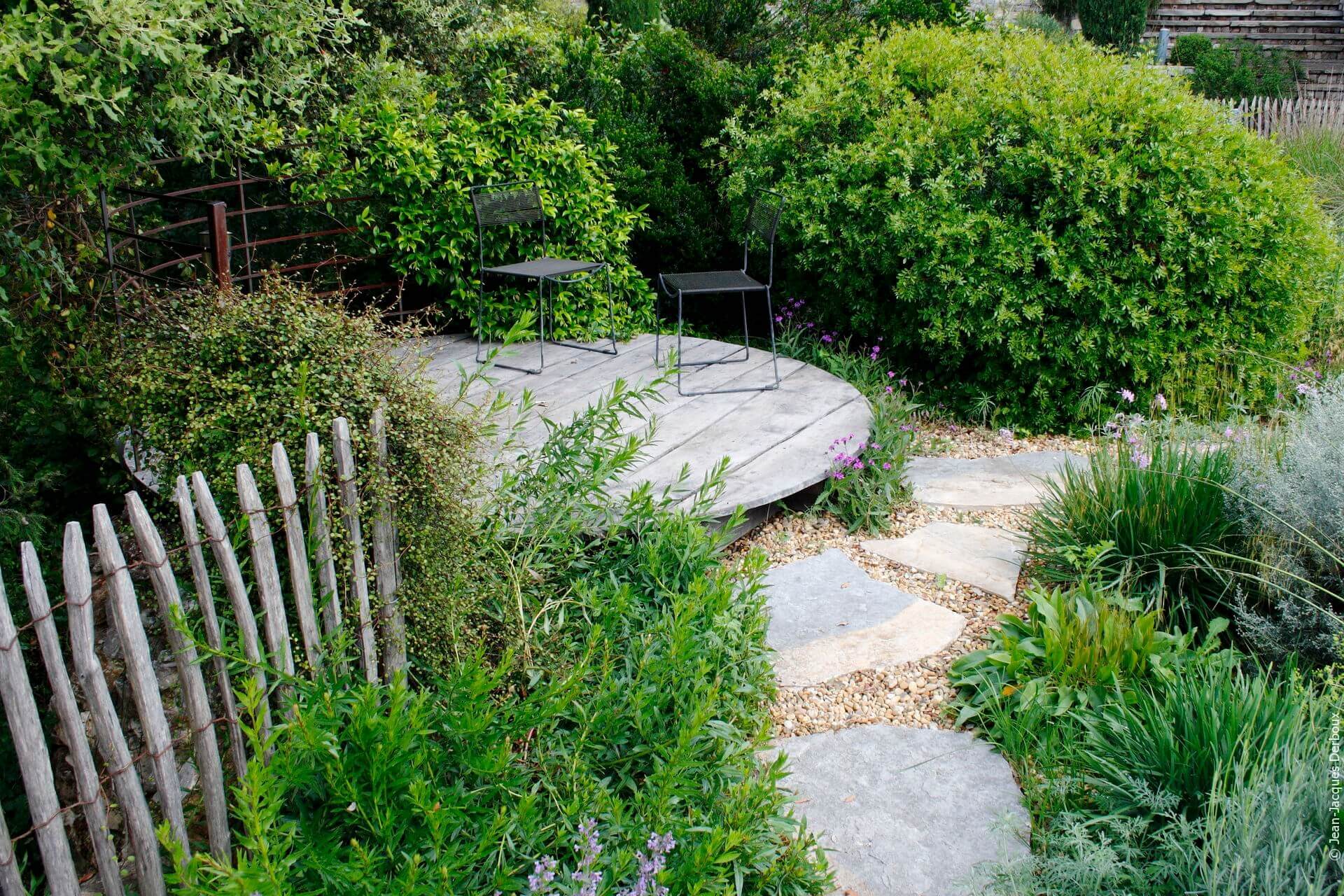 Chaise en métal, terrasse ronde en bois, dalle en pierre, gravier, ganivelle en chataignier, garde corps en métal, plantes pour jardin sec.